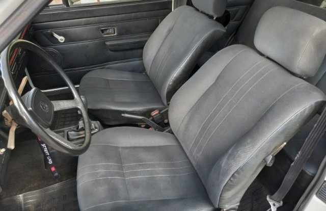 Foto tirada de lado mostrando os bancos dianteiros do carro que são em tecido preto com costuras em cinza claro.