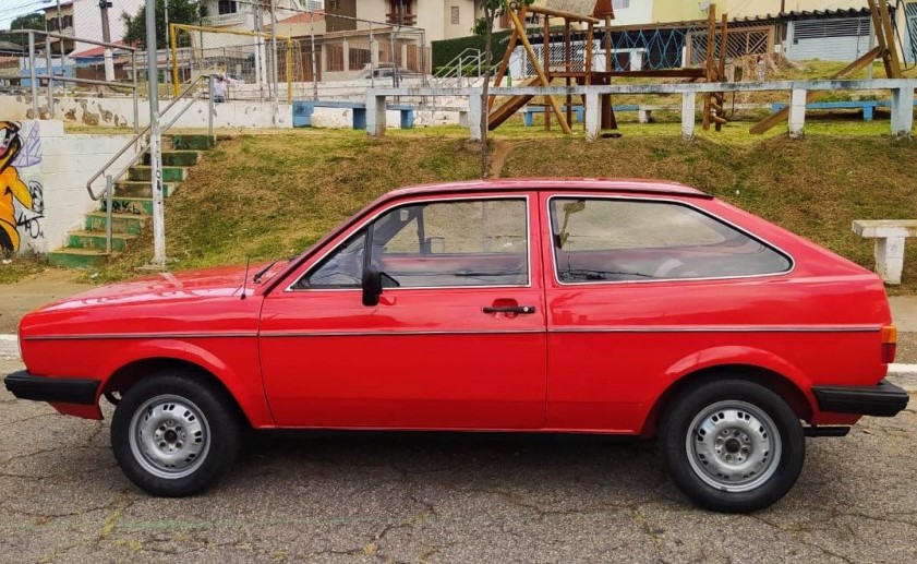 Foto mostrando a lateral esquerda de um Volkswagen Gol G1 vermelho estacionado em uma rua de asfalto.